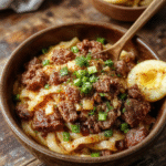 A vibrant bowl of Mongolian ground beef noodles featuring glossy ground beef, colorful julienned vegetables, and tender noodles garnished with chopped green onions and sesame seeds, styled on a rustic wooden table with a rich sauce coating each ingredient.