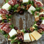 A vibrant, circular charcuterie wreath featuring a variety of colorful cheeses, cured meats, fresh fruits, nuts, and herbs. The arrangement is on a rustic wooden board, with textures ranging from creamy cheeses to shiny cured meats, accented by sprigs of fresh herbs and small bowls of spreads. The presentation is festive and inviting, perfect for holiday gatherings, with a backdrop of seasonal decor and warm lighting highlighting the assortment's freshness and abundance.