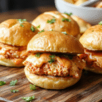 Three golden-brown mini slider buns filled with shredded buffalo chicken topped with blue cheese crumbles and pickled jalapeños, served on a wooden board with extra sauce drizzle and fresh cilantro, vibrant red-orange hues contrasted with white cheese and green garnish