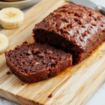 A glossy, slice of rich chocolate banana bread resting on a wooden cutting board, showing a soft, moister crumb with visible banana strands and melted chocolate pockets, dusted with powdered sugar, with a drizzle of chocolate sauce and sliced banana on the side, natural daylight, soft shadows, shallow depth of field